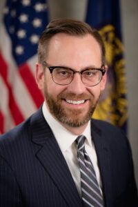 Portrait of a person wearing a dark suit, white shirt, and striped tie, standing in front of U.S. and Nebraska flags.